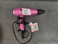 Pink Proliss hair dryer placed on gray surface, showing front nozzle and body with cord wrapped around handle.