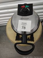 Top view of the Hamilton Beach waffle machine showing stainless steel exterior, control knob with power and ready indicator lights, and handle.