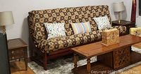 Futon set up as a sofa with patterned cushions and three pillows on a rug between wooden side tables with lamps and coffee table in front.
