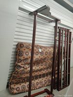 Disassembled wooden bed frame components alongside a patterned futon mattress, showing frame parts standing upright and the mattress leaning against the wall.