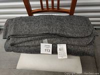 Folded gray polyester fleece blanket displayed on a white cushioned chair with a wooden backrest. A label and a tag with printed information are placed on top, showing details and lot number 113.