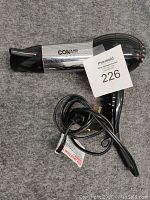 Photo of black and silver Conair hair dryer with concentrator nozzle and power cord coiled beside it on a grey fabric surface with a tag number 226. Shows side and handle details.
