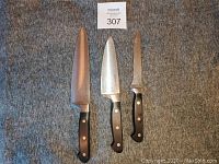 Photo shows 3 Faberware kitchen cutting knives with black handles and metal blades laid out on carpeted floor with lot number card above.
