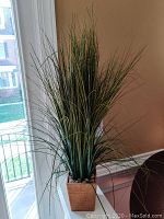 Artificial lemongrass plant with green blades in a wooden square pot with decorative stones at the base, displayed indoors near a window.
