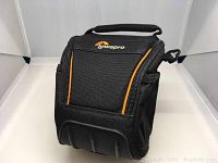 Front angled view of black Lowepro camera bag with orange accents, showing front zip and handle.