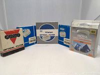 Five camera filter boxes from brands COZO, MONAMI (two filters), HOYA, and M Vision, all 49 mm size, arranged side-by-side against white background, showing brand names and packaging.