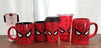Five Spider-Man themed drinkware items on wooden surface: two red ceramic mugs, one red plastic travel mug with black lid, one clear red-tinted cold drink cup, one red ceramic stein mug. All have black spider web patterns and Spider-Man eye designs.
