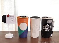 Front view of three Starbucks collectible travel mugs, showing their distinctive designs and black plastic lids.