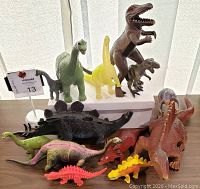Photo of 13 toy dinosaurs in various colors and sizes, arranged on a white surface in front of vertical blinds.