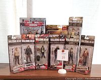 Full lot of 10 Todd McFarlane Walking Dead collectible action figures unopened in packaging, displayed standing on wooden surface in front of light curtain.