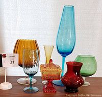 Front view of seven colorful glass items clustered on shelf: tall light blue vase, large amber goblet, green and red small vases, yellow slender vase, blue wine glass and orange covered dish. No visible damages.