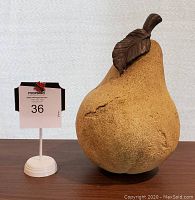 Front view of textured pear sculpture next to lot number stand for scale showing approximately 10 inches tall with brown leaf and stem detail.