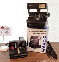 Two vintage black Polaroid OneStep instant cameras, one with original box, displayed with auction sign