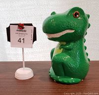 Front view showing the green dinosaur with textured scales, large orange-ringed eyes, and sitting posture next to a number stand.