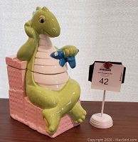 Front view of the dinosaur cookie jar showing full seated green dinosaur holding a blue fish, sitting on a pink chair.