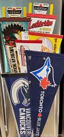 Photo showing six folded vintage banner pennants stacked with packaging. Includes a visible Indian head headdress theme, Lethbridge Alberta travel pennant, and blue sports pennants for Vancouver Canucks and Toronto Blue Jays.