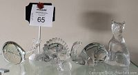 Six vintage clear art glass animal figurines arranged on glass shelf, including brontosaurus, stegosaurus, whale, cat, and hedgehog; good visibility of overall condition and some flaws