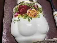 Close-up of white bone china bowl with red and yellow rose floral pattern and green leaves, scalloped edges with gold trim.