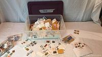 Plastic storage tub filled with loose and bagged postage stamps, some on paper pieces.