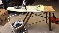 Antique wooden ironing board with replaced fabric cover, electric iron on top, folded step stool with hairdryer resting on it.