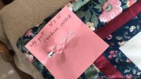 Close-up of pink note describing the quilt as 32 inch by 32 inch, hand quilted (machine stitched) with a safety pin attached, placed on a tan textured surface beside a beige knitted item.