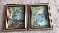 Photo of two original framed oil paintings showing landscape scenery side by side on a light-colored textured surface