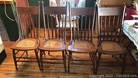 Six antique wooden chairs with turned spindles, carved back panels, and cane woven seats on wood floor.