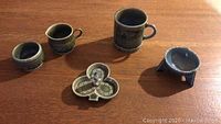 Overview of 5 pottery pieces on wood surface including mug, creamer, small bowl, trinket holder and tripod stand.