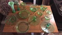 Wide view of the 13-piece green depression glass collection displayed on wooden table, showing variety of plates, bowls, candy dish, candle holders, and two vases.