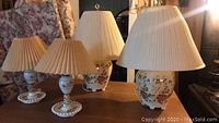 Four vintage ceramic lamps on wooden table with floral-patterned upholstery behind; two smaller white painted lamps with pleated beige shades and two larger floral decorative lamps with cream pleated shades.
