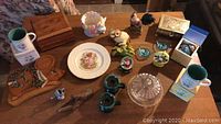 Photo shows 19 assorted decor items including figurines, mugs, wooden box, plates and glassware on a wooden table.