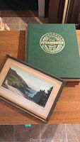 Green hardcover atlas with 'Atlas of Peterborough County' embossed on cover, alongside framed coastal artwork.