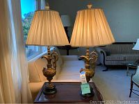 Pair of ornamental goldtone table lamps with cream pleated lampshades on a wooden side table in a living room setting.