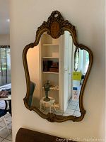 Full view of vintage wall mirror hanging on wall showing curving gold-toned carved frame and reflection of room.