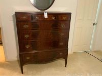 Full view of vintage wooden chest of drawers showing all 7 drawers closed, dark wood finish, brass oval handles, standing on short legs.