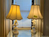 Pair of chalkware cherub lamps on a glass table in front of window with closed blinds and cream curtains.