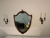 Full view of shield-shaped wood-framed mirror mounted on wall with two brass double candle wall sconces on each side.