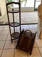 Wooden folding 3-tier shelf table next to wooden box with brass accents on tiled floor near glass doors.
