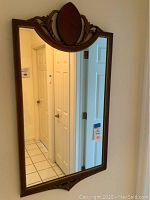 Full view of the vintage wall mirror hanging on a beige wall showing its rectangular shape and red oval top accent with leaf carvings.