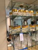 Full view of 9 decorative gold trimmed highball glasses and 6 crystal drinking glasses on glass shelves