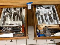 Two open wooden drawers showing assorted stainless steel flatware including forks, spoons, and knives in white trays, alongside cooking and kitchen utensils like knives, cutters, and wooden tools