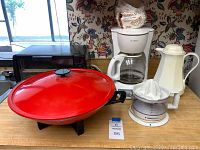 Photo showing red electric wok with lid, white coffee maker with glass carafe, white juicer, and a thermos carafe on a kitchen counter.