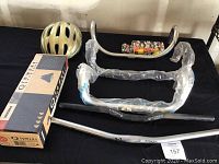 Photo displaying bike accessories including a pale yellow bike helmet, Yakima Q Towers boxed roof rack component, four different silver and black handlebars of various shapes, and a small assortment of bike parts.
