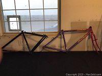 Two bicycle frames side by side leaning against wall, purple/red Specialized Hard Rock frame with fork on right and black Specialized Stumpjumper frame on left