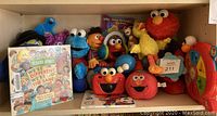 Shelf with Sesame Street toys including stuffed animals of Elmo, Cookie Monster, Big Bird and Ernie, Elmo-themed toy cameras, and a Sesame Street book in front.