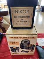 Photo shows a black Nikor stainless steel film developing tank labeled for two 35mm films, sitting on top of a box labeled 'CUT YOUR FILM COST IN HALF ALDEN 74 35 MM BULK FILM DAYLIGHT LOADER' with an illustration of the loader.