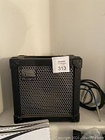 Roland Cube 15 amplifier shown on a shelf with attached cable coiled to the side. Black color with metal grille and Roland logo visible.