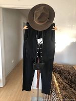 Full view of the outfit hung with hat on top, vest, pants, and leather crossbody bag in front.