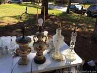 Four table lamps on a white folding table outdoors showing the full set including porcelain, lead crystal, and acrylic bases.