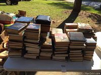 View of lots of stacked books on a white table outdoors beneath a tree, several are hardback and some softcover, various titles and genres.